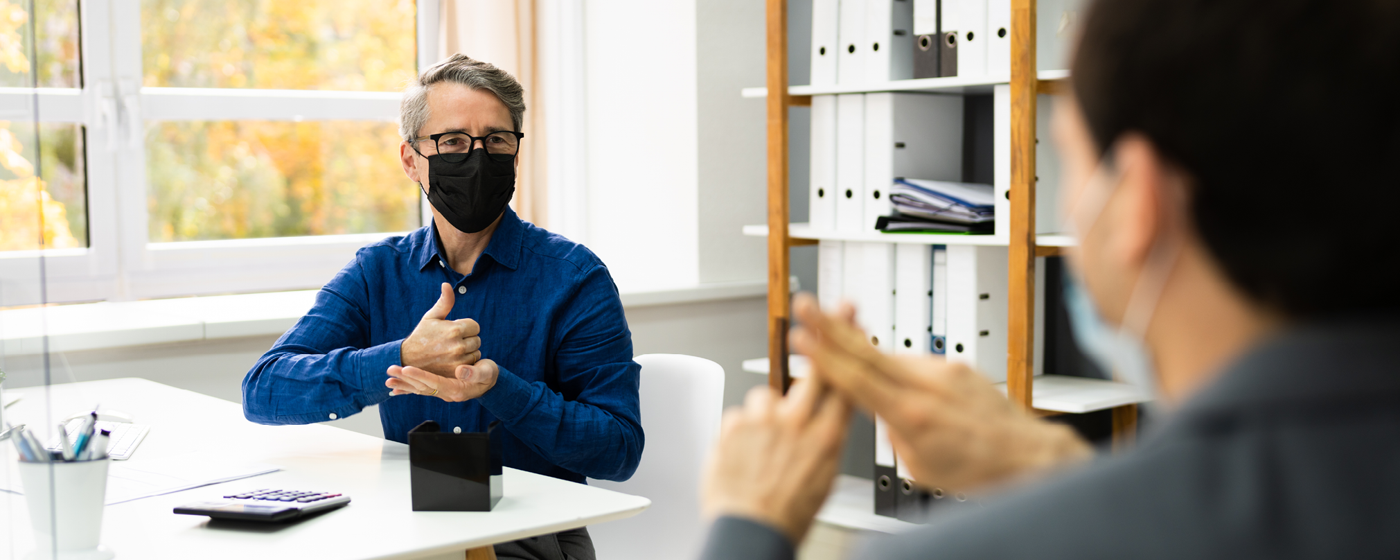 Two people wearing masks, communicating in sign language in an office.