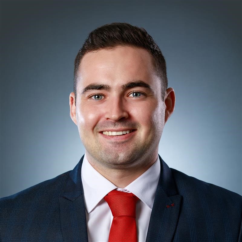Person in a suit with a red tie smiling against a blue background.