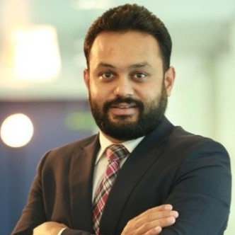 Smiling person with a beard in a suit and checkered tie, standing with arms crossed in an office setting.
