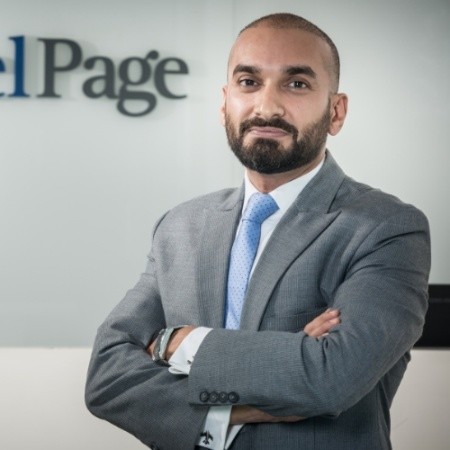 Person in a suit with arms crossed, standing in front of a logo.