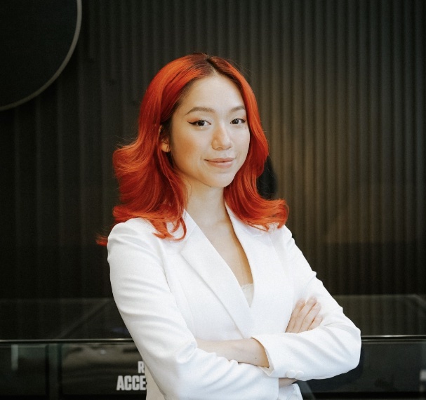 Person with red hair in a white outfit stands confidently with arms crossed.