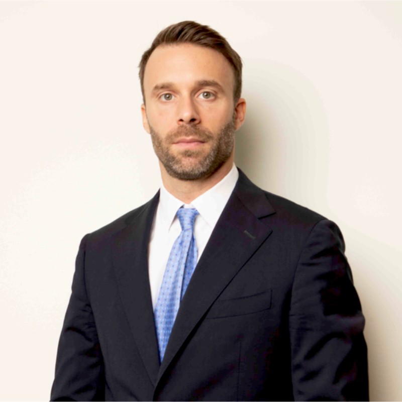 Person in a suit with a blue tie standing against a neutral background.