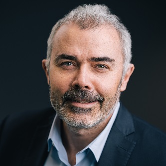 Portrait of a smiling person with short gray hair and a beard, wearing a suit and light shirt.