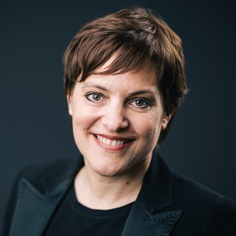 Person smiling with short hair and dark suit against a dark background.