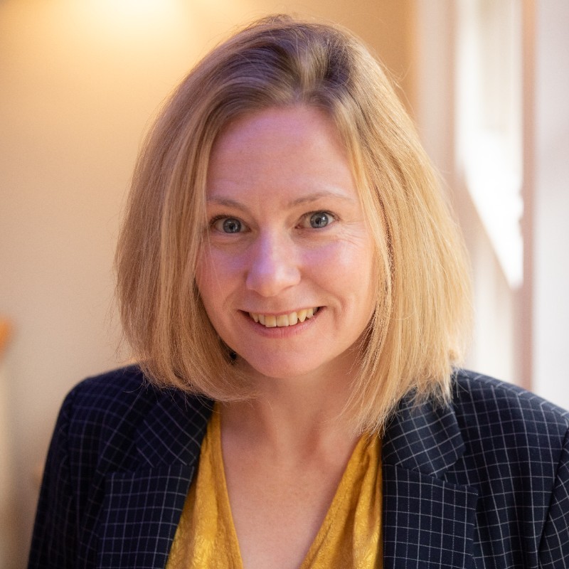 Person smiling indoors in a bright space, wearing a patterned blazer and yellow top.