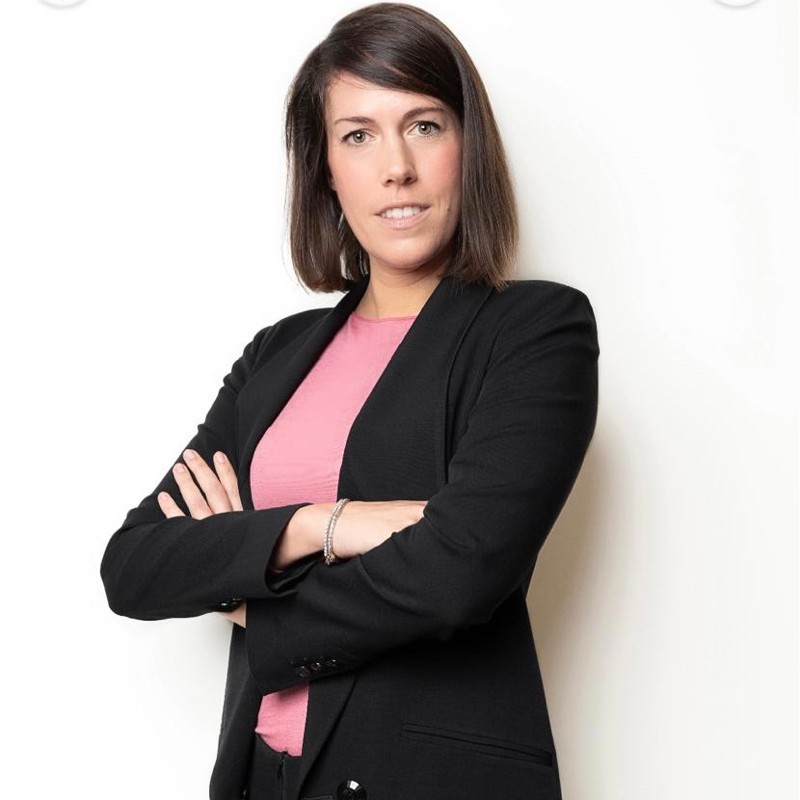 Person in a black suit with arms crossed, standing against a white background.