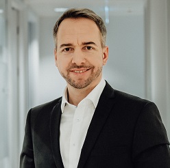 Smiling person in a suit standing in a bright hallway.