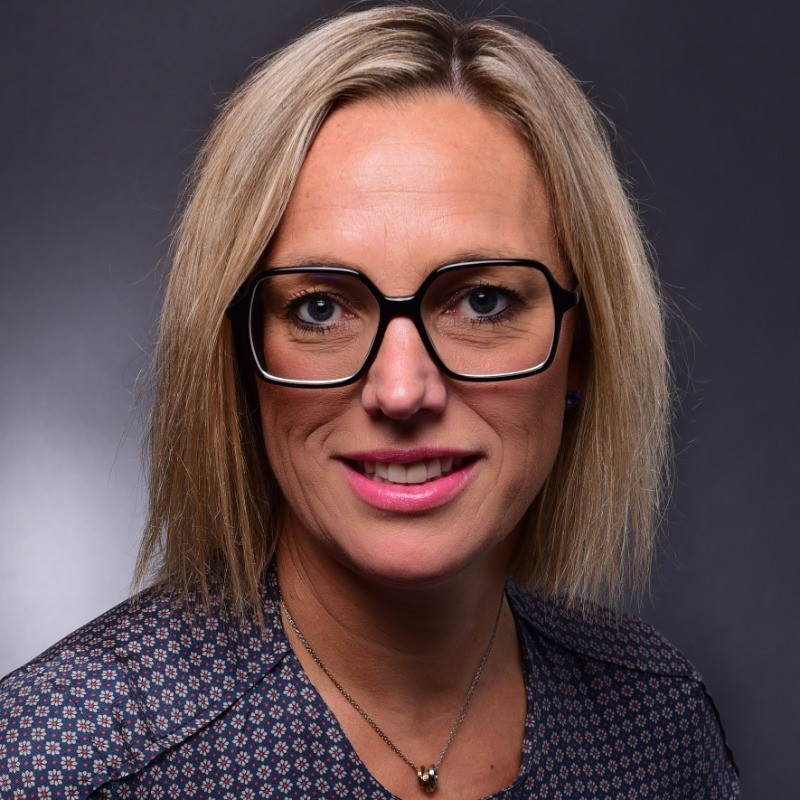 Person with short hair and glasses, wearing a patterned shirt, smiling against a dark background.