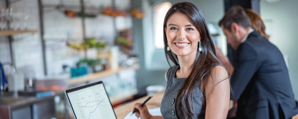 Person smiling at camera while holding a pen, with a laptop displaying graphs in an office setting.