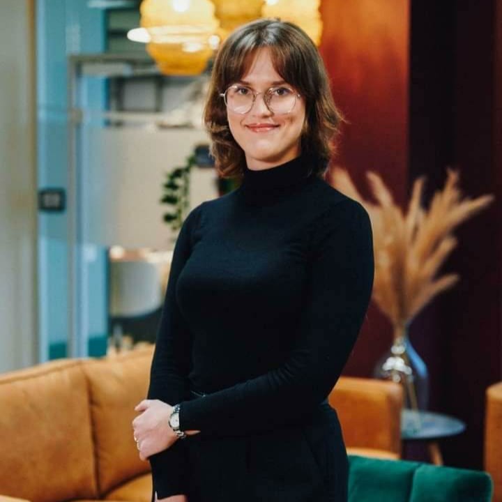 Person wearing glasses and a black outfit stands in a cozy room with warm lighting.