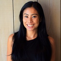 Person smiling, wearing a sleeveless black top, standing against a wooden background.