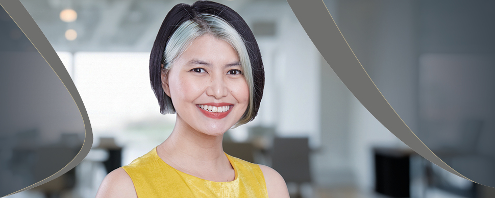 Person smiling with short hair, wearing a yellow top, in a blurred office setting.