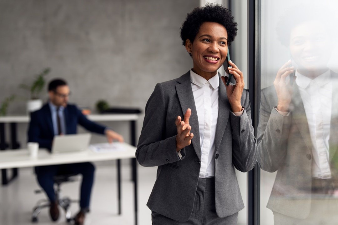 Person in a suit smiling while on the phone near a window at the office.