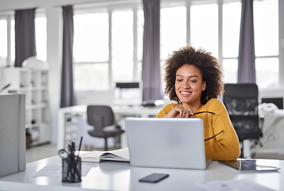 Person smiling while working on a laptop in a bright office.