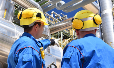 Two industrial workers wearing blue protective clothing, yellow helmets, and ear protection, inspecting large metal pipes and equipment at an outdoor facility.