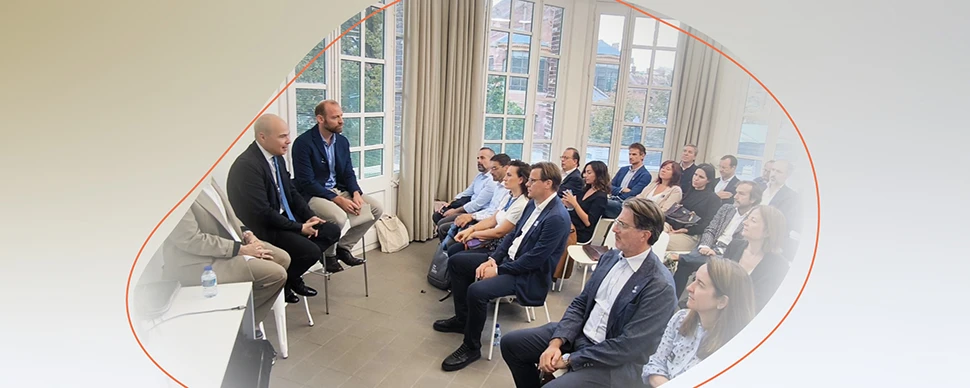 Group attending a discussion in a bright room, with several people seated in rows listening to a small panel at the front.