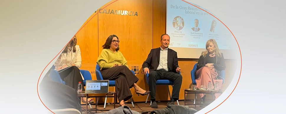 Panel of speakers seated on stage during an event, with a presentation screen and wooden backdrop behind them.