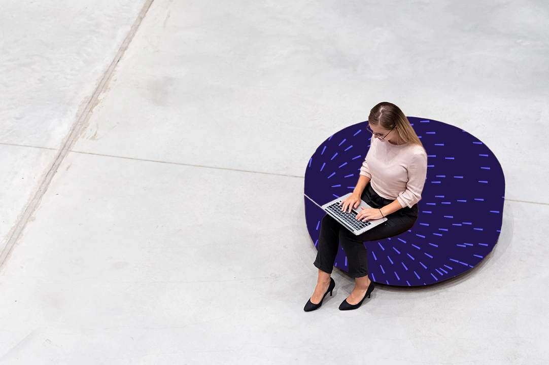 Person using a laptop while seated on a circular bench in an open indoor space.