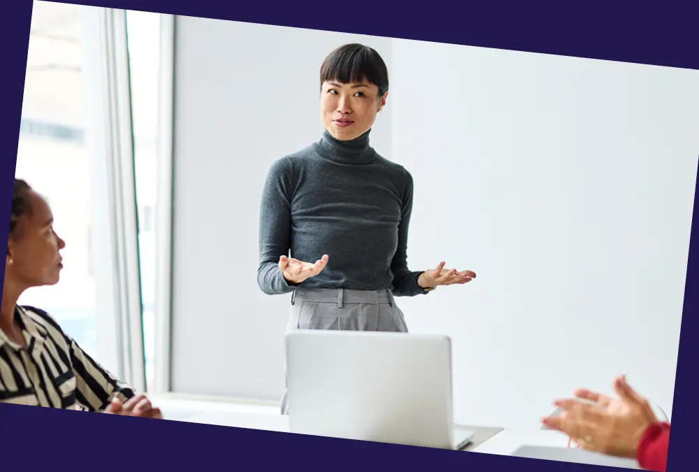 Person presenting to colleagues during a meeting, standing beside a laptop in a bright office.