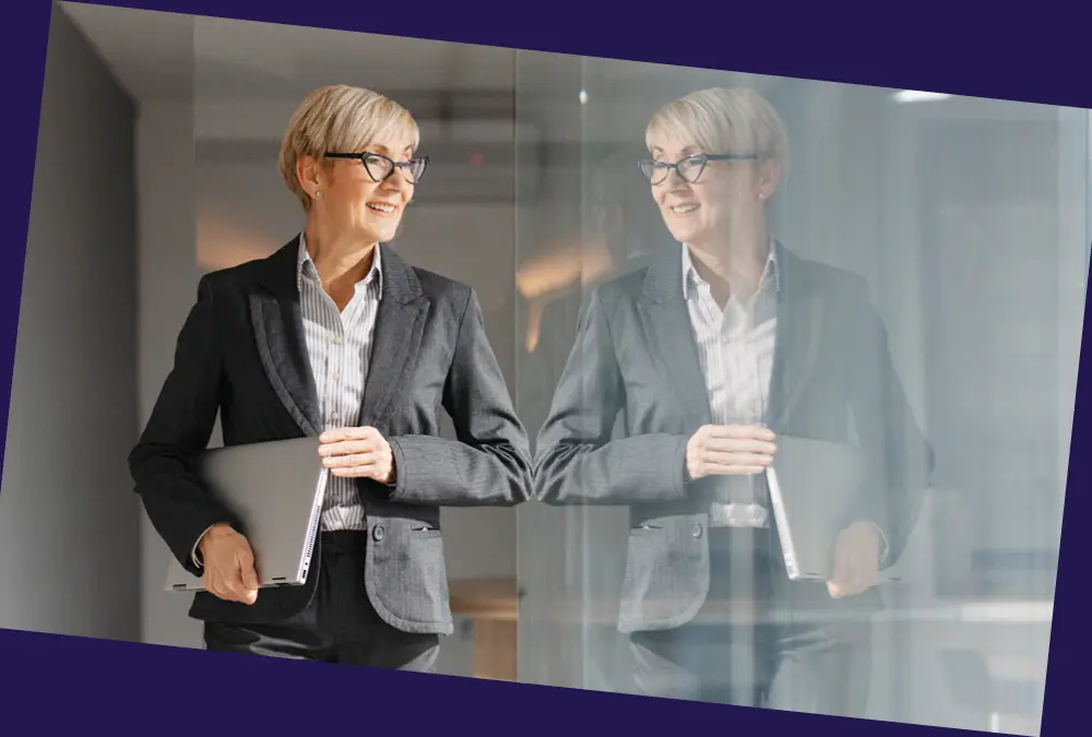 Person in business attire holding a laptop while standing beside a glass wall in an office.
