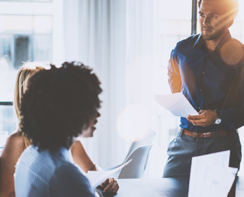 People in an office meeting, discussing documents in bright sunlight.