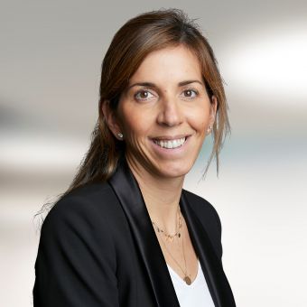 A person smiling, wearing a black blazer and white shirt, blurred background.