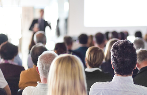 A large audience listens to a presentation in a bright conference room.