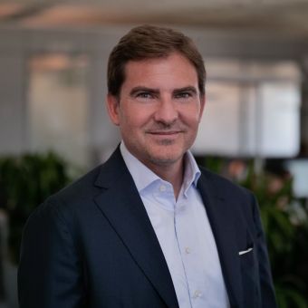 Person in a suit smiling indoors with plants in the background.