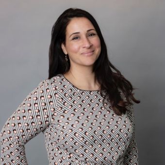 Person smiling in front of a gray background, wearing a patterned top.
