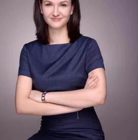 Person in a blue dress with arms crossed, smiling slightly.