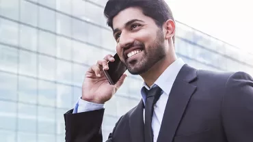 Person in a suit smiling while talking on a phone in front of a glass building.