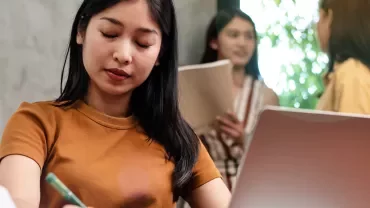 Person focuses on writing at a desk, with two others talking in the background.