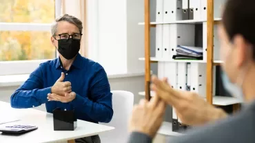Two people wearing masks, communicating in sign language in an office.
