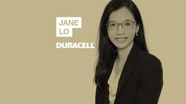Person in a formal outfit with glasses, smiling against a solid background.