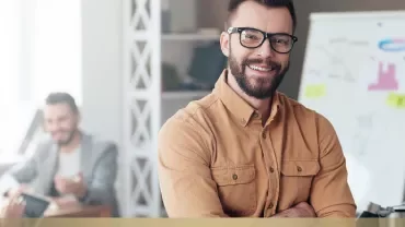 Person smiling, standing in an office with a whiteboard and two people in the background.