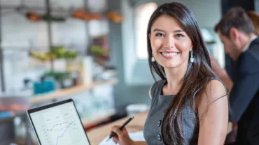 Person smiling at camera while holding a pen, with a laptop displaying graphs in an office setting.