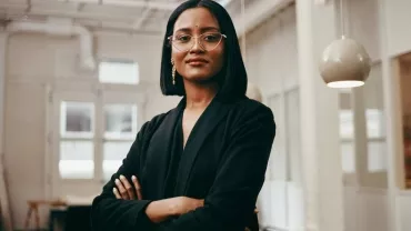 Person in a black blazer with arms crossed, standing confidently indoors.