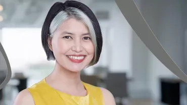 Person smiling with short hair, wearing a yellow top, in a blurred office setting.