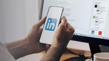 Hands holding a smartphone displaying the LinkedIn logo, with a computer screen in the background.