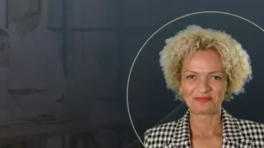A professional portrait of a woman with curly blonde hair, wearing a patterned blazer, overlaid on a dark background with subtle graphic lines and the faint image of two women working on a laptop in the background.