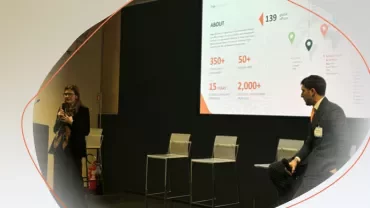 Two people participating in a presentation, with one speaking on stage and a large slide displayed behind them.