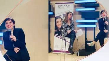 Three individuals seated on a stage during a discussion, with microphones and a banner displayed in the background.