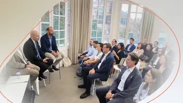 Group attending a discussion in a bright room, with several people seated in rows listening to a small panel at the front.