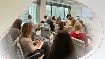 Group attending a presentation in a modern meeting room, with a speaker standing at the front near a screen.