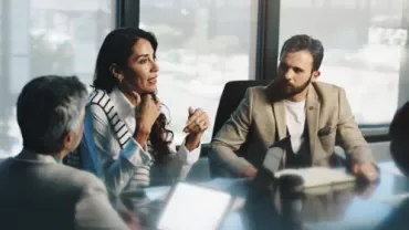 People meeting around a conference table in a modern office, with one person speaking and others listening.