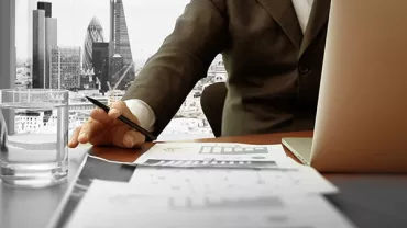 Person in a suit working at a desk with a laptop and papers, cityscape visible through the window.