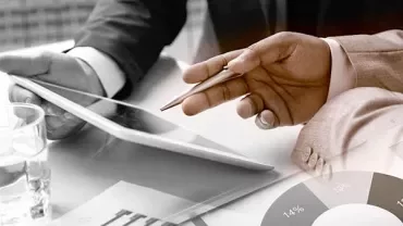 Hands holding a tablet and pen above paperwork with pie charts and graphs.
