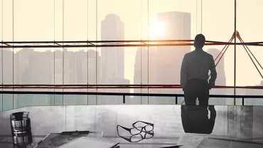 Person looking at city skyline through large office window, desk in foreground.