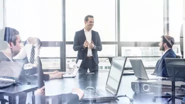Person leading a business meeting in a modern office with several people seated around a table.