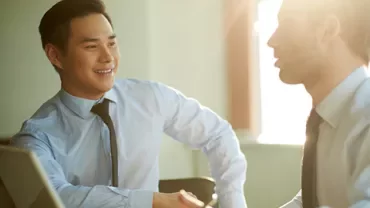 Two people in an office setting discussing at a table with a laptop.